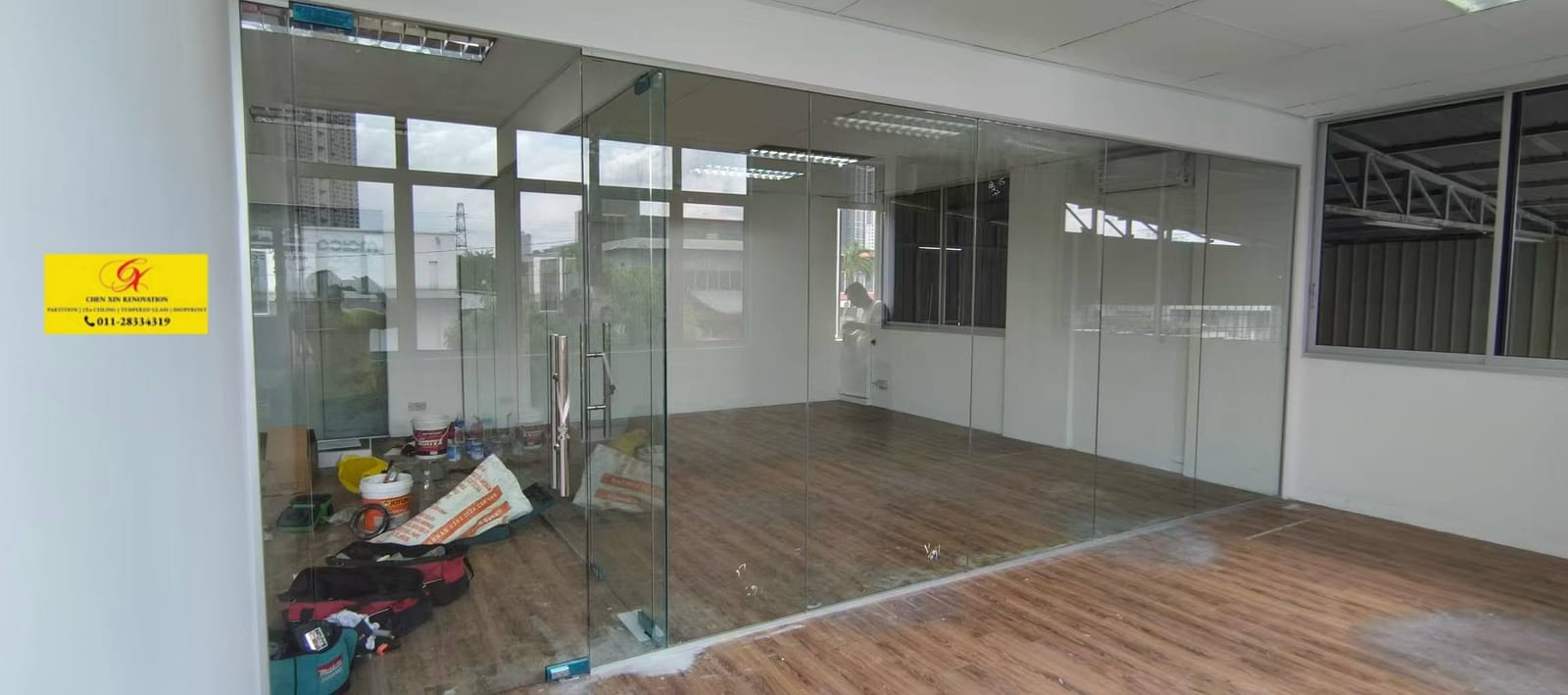 同一间办公室施工中：无框玻璃已经装上地面固定件，地上还放着工具袋和水泥袋，天花尚未封顶。